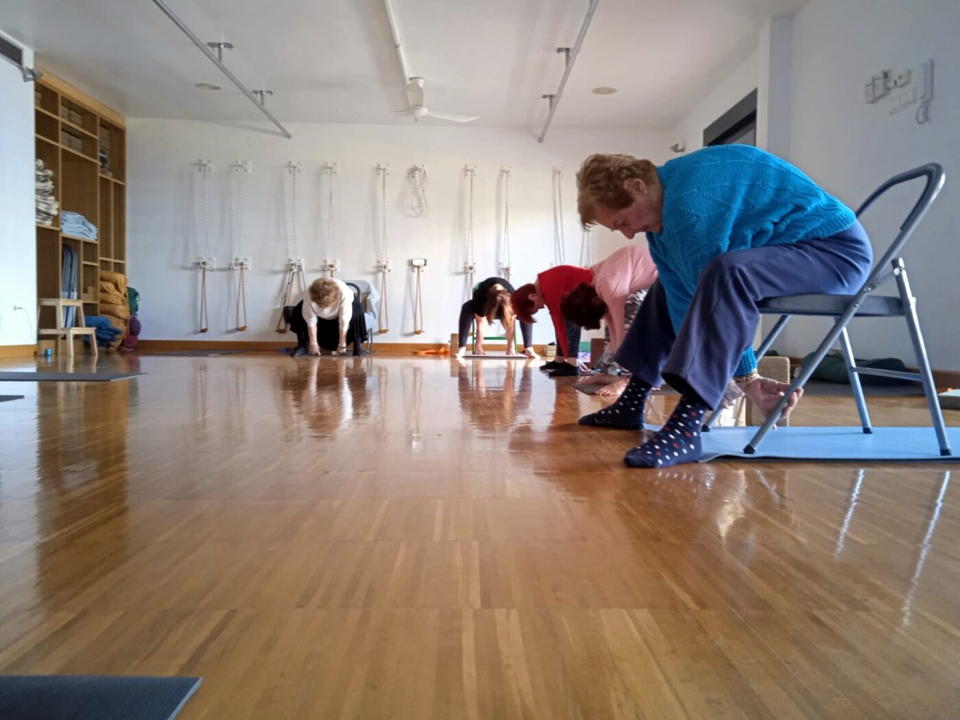 Practicante de yoga adaptado apoyada en una silla, en una flexión hacia delante, con compañeras practicando al fondo