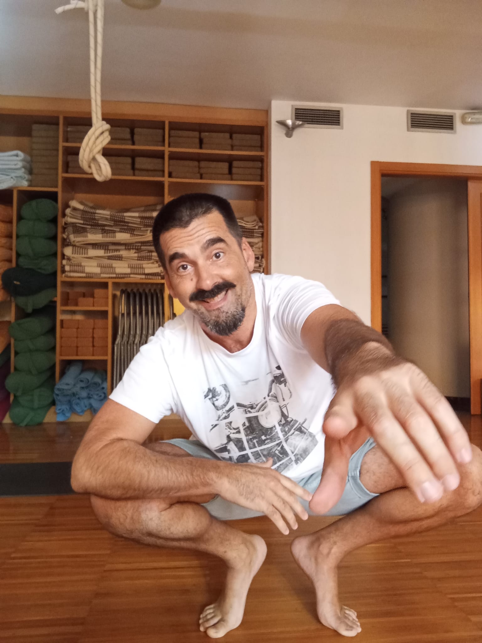 Francis sonriendo en cuclillas en la sala de yoga, señalando a cámara